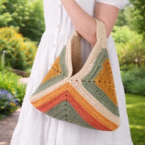 May include: A handmade crochet bag with a geometric design. The bag features stripes of orange, green, beige, and rust colors. The bag has a matching beige handle and is held by a person wearing a white dress. The background is a blurred garden.