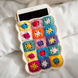 Könnte beinhalten: Eine farbenfrohe, gehäkelte Tablethülle mit Patchwork-Design. Die Hülle besteht aus Quadraten in verschiedenen Farben, darunter Gelb, Blau, Rosa und Orange, mit einem weißen, wellenförmigen Rand. Ein roter Knopf sichert die Oberseite. Ein weißes Tablet befindet sich darin.