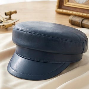 May include: A navy blue leather cap with a rounded crown and a short, stiff peak. The cap features visible stitching and a classic, nautical design. The hat is a solid colour and appears to be made of a smooth material.