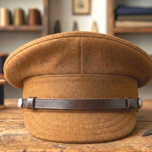 Può includere: Un berretto militare in feltro marrone con corona strutturata e parte superiore piatta. Un cinturino in pelle marrone scuro corre orizzontalmente sulla parte anteriore, fissato da due bottoni metallici argentati su ciascun lato, sopra una visiera curva.