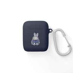 May include: Navy blue AirPod case with a silver carabiner clip. The case features a cartoon illustration of a white rabbit reading a book. The case is designed to protect and carry wireless earbuds.
