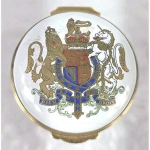 May include: A round gold-toned metal compact with a white enamel top featuring the Royal Coat of Arms of the United Kingdom. The design includes a lion and a unicorn flanking a shield, with the motto DIEU ET MON DROIT and HONI SOIT QUI MAL Y PENSE.