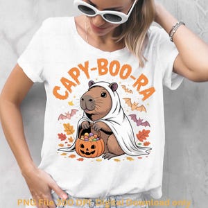 May include: White t-shirt with a Halloween-themed graphic. The design features a capybara dressed as a ghost, holding a pumpkin basket filled with candy. The text "CAPY-BOO-RA" is arched above the capybara in orange.