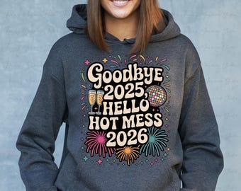 Puede incluir: Una sudadera con capucha gris oscuro con un gráfico colorido que dice "Goodbye 2025, Hello Hot Mess 2026". El diseño incluye copas de champán, una bola de discoteca y fuegos artificiales. Prenda casual y cómoda.