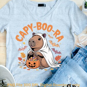May include: Light gray V-neck t-shirt featuring a capybara dressed as a ghost, holding a pumpkin basket filled with candy. The shirt has the text "CAPY-BOO-RA" in orange, with bats and autumn leaves. A fun Halloween design.