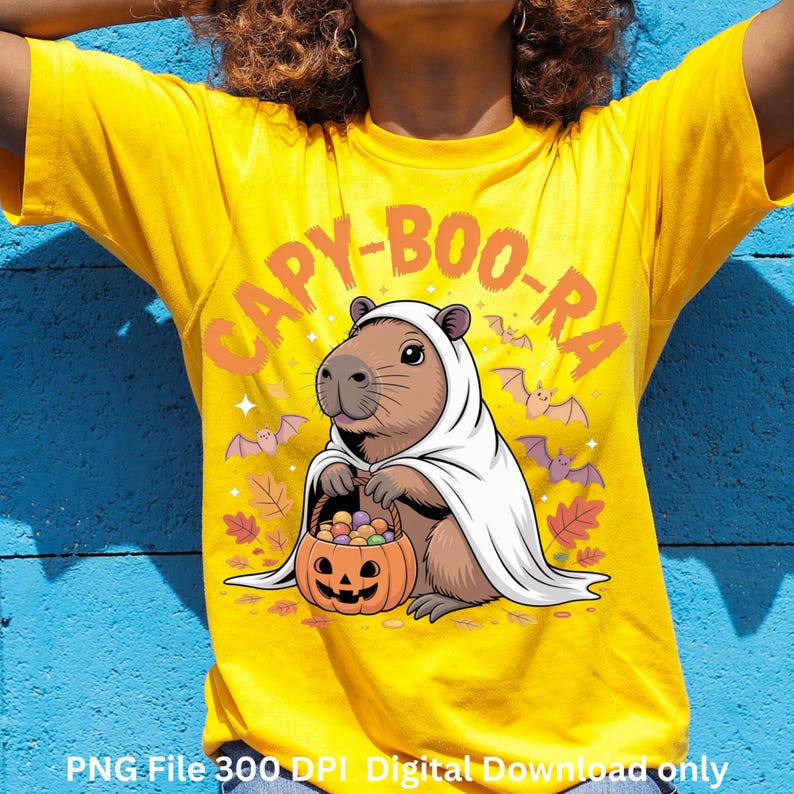 May include: Yellow t-shirt with a Halloween-themed graphic. The design features a capybara in a white ghost costume holding a jack-o'-lantern filled with candy. The text "CAPY-BOO-RA" is arched above the capybara, with bats and leaves around the design.