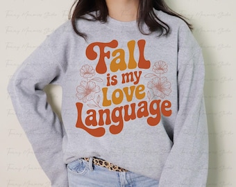 Può includere: Una felpa grigio chiaro con la scritta "Fall is my Love Language" in arancione e giallo. Il design include elementi floreali. È un capo a maniche lunghe.