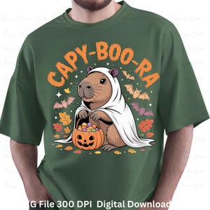 May include: Green t-shirt featuring a Halloween-themed graphic. The design includes a capybara dressed as a ghost, holding a jack-o'-lantern basket. The text "CAPY-BOO-RA" is arched above the capybara, with bats and autumn leaves.