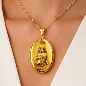 May include: Gold-coloured oval pendant necklace featuring a detailed cat wearing a crown with ruby and emerald accents. The pendant hangs from a delicate chain. The cat pendant is approximately 3.8 cm in height.