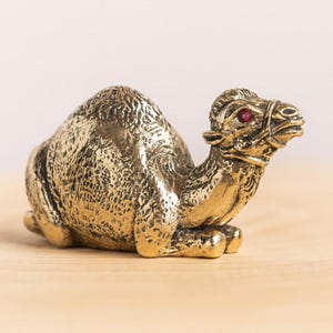 May include: A detailed brass camel figurine in a resting position. The camel has a textured surface and a red gem for an eye. The figurine is set on a light-colored wooden surface.