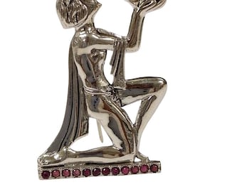 May include: Silver-toned brooch depicting a kneeling figure holding an object aloft. The figure is kneeling on a rectangular base adorned with small, red gemstones. The brooch has a polished finish and is set against a white background.