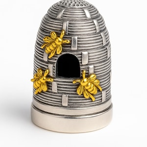 Puede incluir: Un dedal plateado con forma de colmena, adornado con tres abejas doradas. El dedal tiene una pequeña abertura y está diseñado con líneas horizontales y detalles rectangulares.