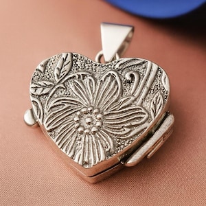 Op de afbeelding: Zilveren hartvormige locket met een bloemenontwerp. De locket is gedetailleerd met een reliëf bloem- en bladerpatroon. Het heeft een scharnierende opening en een driehoekige beugel voor een ketting.