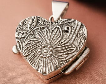 May include: Silver heart-shaped locket with a floral design. The locket is detailed with an embossed flower and leaf pattern. It has a hinged opening and a triangular bail for a chain.