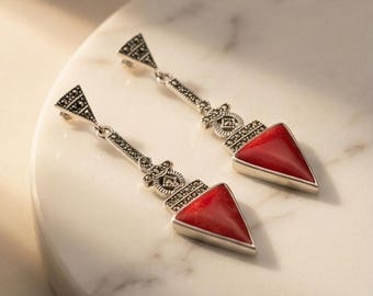 May include: A pair of silver-toned drop earrings featuring red triangular stones. The earrings have a geometric design with small, dark accents and a triangular top. The red stones are set in a silver frame.