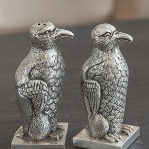 Pode incluir: Um par de saleiros e pimenteiros prateados em forma de pássaro. Os saleiros e pimenteiros são detalhados com penas texturizadas e assentam em bases quadradas. Um tem orifícios para pimenta.