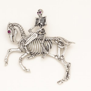 Puede incluir: Broche plateado que representa un esqueleto montando un caballo esquelético. El esqueleto y el caballo tienen detalles intrincados, con ojos de piedras preciosas rojas. Un accesorio único de temática gótica.