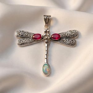 Puede incluir: Colgante de plata con forma de libélula con alas detalladas. Las alas tienen patrones intrincados. El colgante presenta dos piedras preciosas ovaladas de color rojo intenso y una piedra preciosa iridiscente ovalada en la parte inferior.