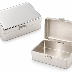 Pode incluir: Uma caixa retangular de metal em tom prateado com um acabamento exterior liso e polido. A caixa é apresentada em dois estados: uma fechada com um mecanismo de fecho simples na frente, e outra aberta, revelando um interior metálico escovado.