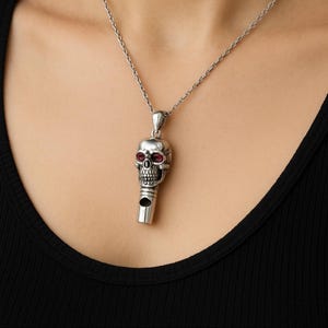 May include: A silver-colored skull pendant with red gemstone eyes hangs from a delicate chain. The skull has detailed features and a whistle-like design. The pendant is worn against a black top.