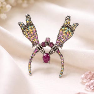 Puede incluir: Broche plateado con un diseño único. Presenta dos formas estilizadas en forma de mano con alas coloridas estilo vidriera. Un gran rubí ovalado está engastado en el centro, con rubíes más pequeños encima. El broche está sobre un fondo blanco.