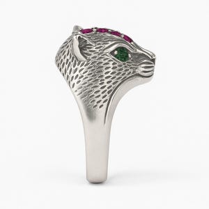 May include: A silver ring featuring a detailed leopard head design. The leopard's head is adorned with small, red gemstones along the top and a single green gemstone for the eye. The ring has a polished finish.