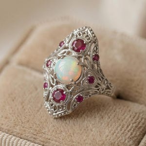 May include: Ornate silver ring featuring a large, round opal center stone and smaller ruby accents. The ring has an intricate filigree design and is set against a soft, neutral background. A vintage-inspired piece of jewelry.