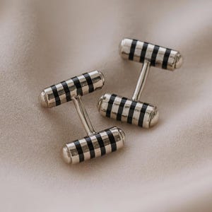 May include: A pair of silver cufflinks with a black and silver striped design. The cufflinks have a T-bar shape and are set against a neutral background. The design is a classic and elegant accessory.