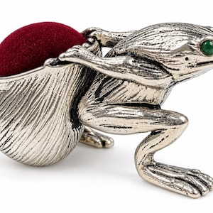 May include: A silver-toned metal frog figurine featuring a textured finish and a single green eye. The frog is depicted in a crouching position, supporting a rounded, deep red velvet pincushion on its back, which is integrated into the design.