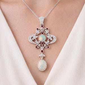 Könnte beinhalten: Eine silberne Anhängerkette mit einem verzierten Design. Der Anhänger hat einen großen ovalen Opal unten, einen zentralen Opal und kleine rote Edelsteine. Das Design umfasst florale und scrollartige Details und ist mit kleinen, klaren Edelsteinen besetzt.