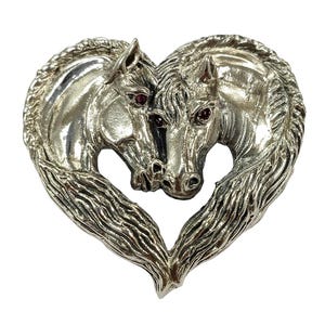 May include: A silver-toned brooch in the shape of a heart, featuring two horses facing each other. The horses have detailed manes and red gemstone eyes. The brooch is likely made of metal and has a polished finish.