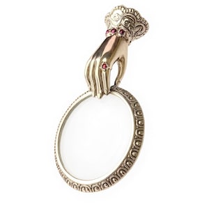 May include: A silver-coloured magnifying glass with an ornate handle shaped like a hand. The hand is decorated with red gemstones and holds a round, clear lens. The lens frame has a decorative pattern.