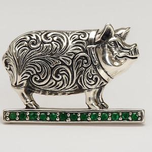 Puede incluir: Figurilla de cerdo plateado con intrincados detalles de volutas. El cerdo se alza sobre una base rectangular adornada con una hilera de piedras preciosas verdes. Es un objeto decorativo, probablemente de colección o una obra de arte. La base mide unos 5 cm.