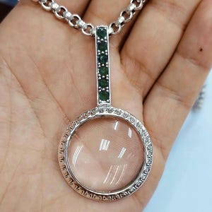 Peut inclure: Un collier en argent avec un pendentif en forme de loupe. Le pendentif est entouré d'une bordure décorative et a un accent de pierre verte sur la chaîne.