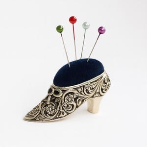 May include: A silver-coloured, shoe-shaped pin cushion with a dark blue velvet top. The shoe features detailed floral and scrollwork patterns. Four pins with coloured heads are inserted into the cushion.