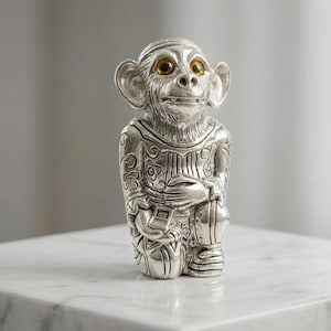 May include: A silver monkey figurine with intricate carvings and yellow eyes. The monkey is in a seated pose, holding an object. The figurine features a decorative pattern on its clothing and is placed on a white surface.