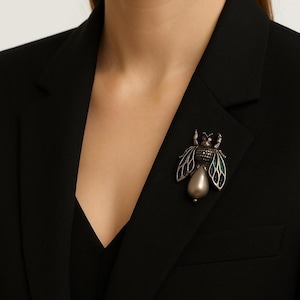 Puede incluir: Broche de abeja plateado con una gota de perla en forma de pera. El broche presenta alas detalladas con detalles de esmalte azul y verde y pequeñas piedras preciosas moradas y transparentes. El broche está prendido a una blazer negra.