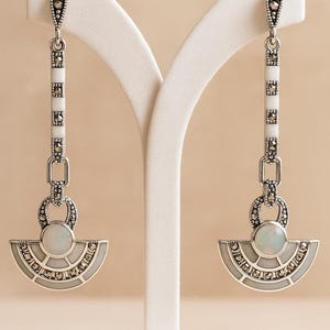 May include: Elegant silver-tone earrings with a white and marcasite design. The earrings feature a fan-shaped bottom with a central opal-like stone and a vertical white and marcasite bar. The earrings are displayed on a white stand.