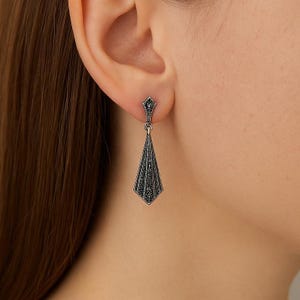 Könnte beinhalten: Nahaufnahme eines silberfarbenen Ohrrings mit geometrischem, vom Art Déco inspiriertem Design. Der Ohrring hat eine rautenförmige Oberseite und einen langen, spitz zulaufenden Anhänger, verziert mit kleinen, dunklen Steinen.