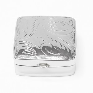 Może przedstawiać: Kwadratowe, srebrne pudełko z otwieraną pokrywą. Pokrywa zdobiona jest misternym, grawerowanym wzorem kwiatowym. Pudełko ma błyszczącą, odbijającą światło powierzchnię i małe zapięcie.