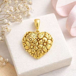 Peut inclure: Un pendentif doré en forme de cœur avec un motif de coccinelle et de fleurs. Le pendentif présente un relief détaillé de fleurs, de feuilles et d'une coccinelle. Le pendentif est présenté sur une surface blanche avec un ruban rose en arrière-plan.