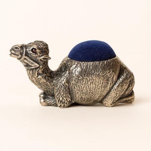May include: A silver-colored camel pin cushion with a dark blue cushion on its back. The camel has a detailed texture and a red gem for an eye. The pin cushion is designed to hold sewing needles and pins.