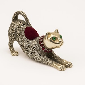 Puede incluir: Un cojín para alfileres con forma de gato plateado con intrincados adornos florales. El gato tiene ojos verdes y un collar con gemas rojas. Un pequeño cojín redondo de color burdeos está en su espalda.
