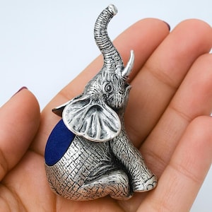 Puede incluir: Una figura de elefante plateada con la trompa levantada. El elefante tiene una piel texturizada detallada y orejas grandes. Un cojín azul está en su espalda. La figura es un objeto decorativo.