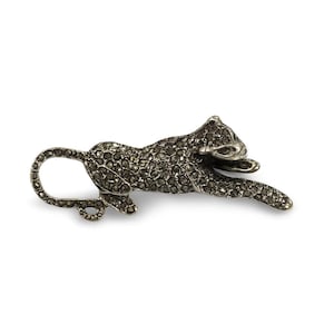 Puede incluir: Broche de metal plateado con forma de leopardo estilizado, tumbado con la cola enroscada. El cuerpo del leopardo está cubierto de pequeñas piedras oscuras, lo que le da un aspecto texturizado. El broche tiene un cierre en la parte posterior.