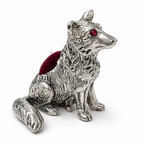 May include: A silver-coloured dog-shaped pin cushion with red gemstone eyes and a dark red cushion. The dog is sitting with its tail curled and has detailed fur texture. A functional and decorative sewing accessory.