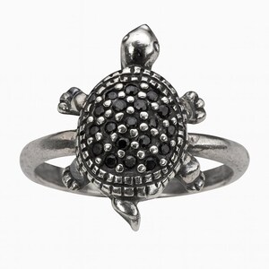 May include: A silver-toned ring featuring a turtle design. The turtle's shell is encrusted with small, black gemstones. The ring has a detailed, textured appearance, with the turtle's head and limbs clearly defined.