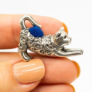 May include: A silver-colored cat-shaped pin cushion with a blue cushion on its back. The cat is in a playful pose, with its front paws extended. The pin cushion is held in a hand with a light orange nail polish.