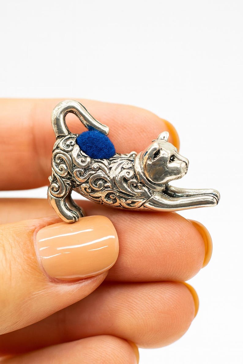 May include: A silver-colored cat-shaped pin cushion with a blue cushion on its back. The cat is in a playful pose, with its front paws extended. The pin cushion is held in a hand with a light orange nail polish.