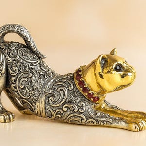 Può includere: Statuetta decorativa a forma di gatto in posizione di allungamento. La testa e le zampe del gatto sono dorate, mentre il corpo è argentato con intricati motivi floreali. Un collare rosso con gioielli adorna il collo. Un oggetto di arredamento unico.